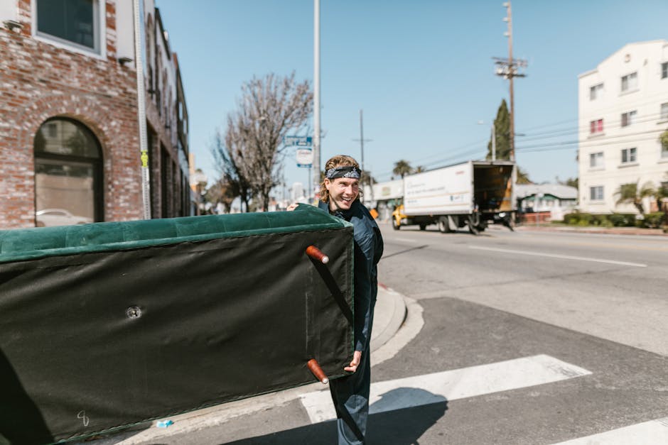 A man with light brown hair wearing a black headband and a dark blue jacket is outside on a street, lifting a large furniture item wrapped in dark protective material with green padding on top, beneath which is visible plastic and fabric wrapping. The furniture is equipped with red handles and is being carried on the pavement near a white crosswalk. In the background, a commercial van and a white truck are parked or moving along the road, with a brick building on the left side and a white residential building on the right. The sky is clear and blue, and a few leafless trees are visible, indicating a cold or winter season. The scene captures part of a home relocation, with the individual involved in moving or furniture transport, reflecting materials, packaging, and loading activities typical of professional removals such as those offered by Man With a Van Lewisham, supporting efficient packing and furniture transport processes.
