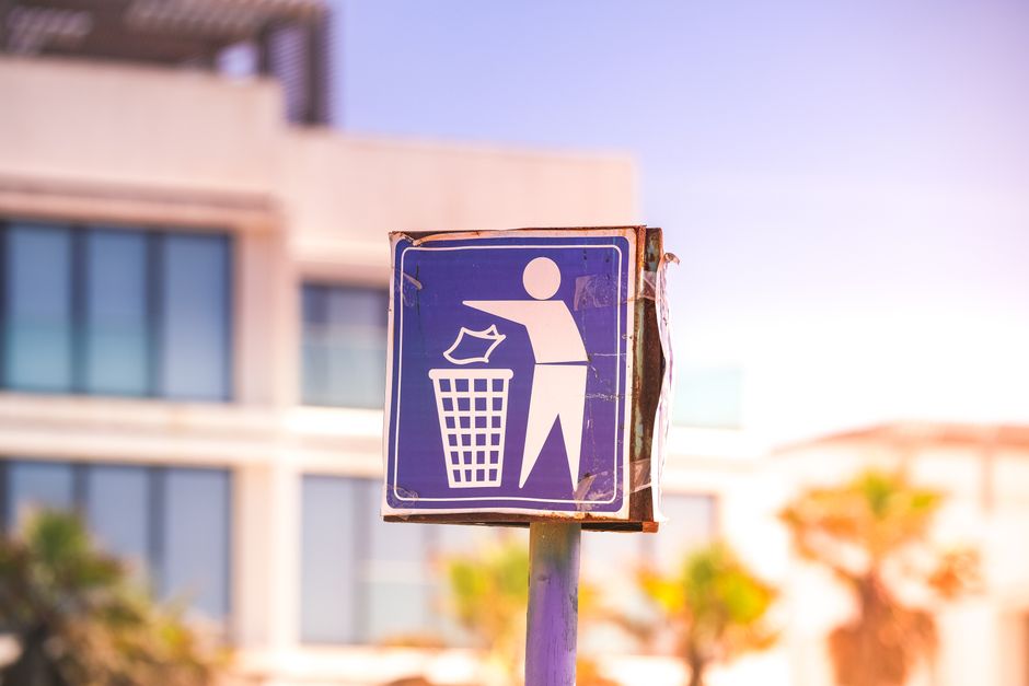 An outdoor scene showing a pile of rubbish and discarded household items near a stone boundary wall under a partly cloudy sky. The waste includes black plastic garbage bags, a yellow plastic storage b