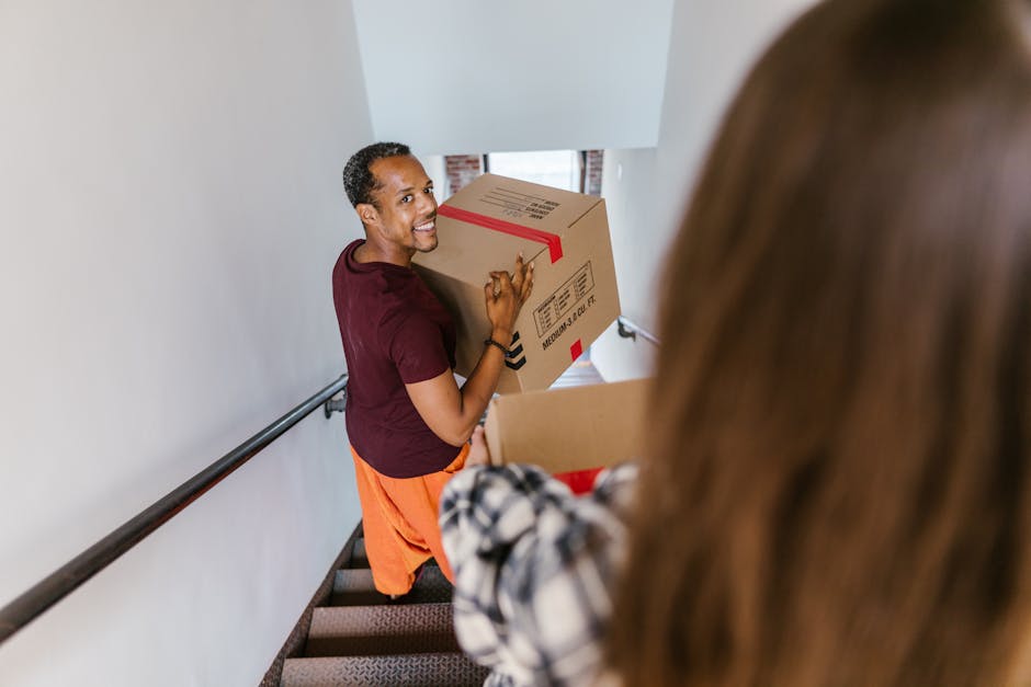 Inside a residential property during a house move, a woman with curly dark hair, wearing a cream-colored sweater, blue jeans, and black shoes, is carefully carrying a medium-sized cardboard box with a